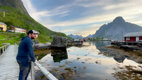 Medium-panning-shot-of-a-person-standing-on-a-wooden-pedestrian-bridge-over-looking-the-famous-lofoten-reine-village-in-Norway,-handheld