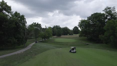 following-golf-carts-driving-on-golf-course,-aerial-drone