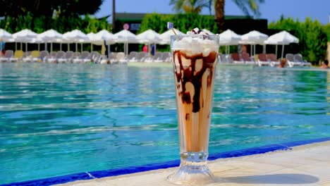 fresh-milkshake-glass-cup-by-calm-sunny-resort-pool-during-summer-vacation,-static-shot