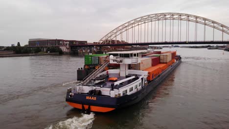 Aerial-View-Off-Stern-Of-Maas-Push-Tow-Barge-Carrying-Cargo-Containers-Along-River-Passing-Approaching-Bridge