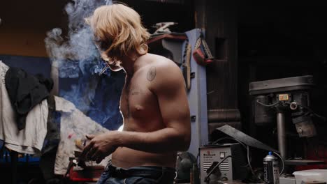 Cinematic-shot-of-mechanic-with-tattoos-lighting-up-a-cigarette-leaning-against-his-workbench