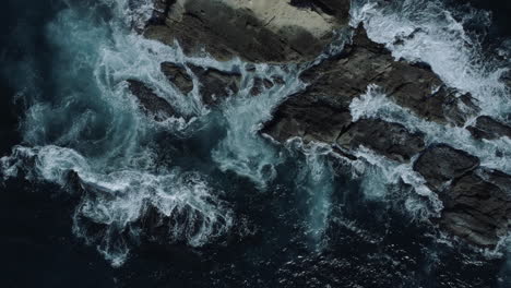 Aerial-shot-from-low-to-high-of-blue-ocean-water-crashing-on-rocks