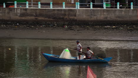 Two-Vietnamese-Fishermen-Rowing-Fishing-Boat-On-Polluted-City-River-In-Vietnam