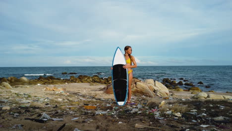 Young-sexy-female-surfer-in-bikini-holding-surfboard-on-polluted-ocean-beach-filled-with-plastic-waste-and-trash,-global-warming-climate-change-concept