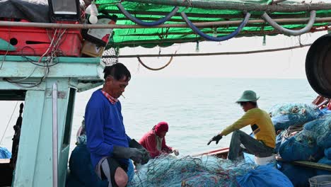 A-fisherman-mending-nets-while-smoking-as-he-takes-the-cigarette-out-of-its-mouth-while-others-also-work-at-the-background,-Pattaya-Fishing-Dock,-Chonburi,-Thailand