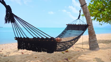 Empty-cotton-rope-net-hammock-with-spreader-bars-lashed-between-two-palm-trees-on-the-sandy-beach-in-front-of-the-turquoise-sea-on-a-tropical-island