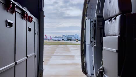Pedestal-down-view-from-inside-airplane-with-opened-door-of-aircrafts-parked-at-London-Gatwick-airport-in-background-on-cloudy-day,-UK