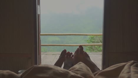 Feet-of-man-and-woman-flirt-together-from-under-blanket-with-the-balcony-and-the-mountain-landscape-background