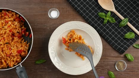 Top-down-shot-of-kitchen-utensil,-spatula-putting-creamy-fusilli-pasta-with-tomato-sauce-on-deep-plate-placed-on-wooden-table-decorated-with-other-spatula,-black-cloth-and-fresh-basil-leaves
