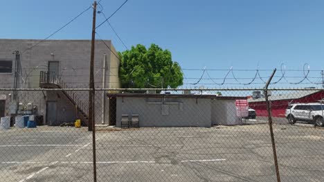 Dolly-down-a-city-alleyway-past-chain-linked-fence-topped-with-barbwire-enclosing-the-back-of-an-industrial-building,-Sunnyslope,-Phoenix,-​Arizona