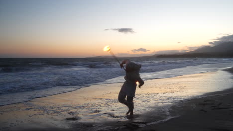 A-man-fire-dancer-in-silhouette-spinning-his-flaming-staff-on-the-beach-at-sunset-with-flames-and-ocean-waves-SLOW-MOTION