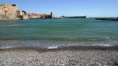 The-historic-old-town-of-Collioure-from-the-pebble-beach-with-gentle-waves-lapping-the-beach-on-a-windy-summer-day