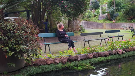 Pretty-pensive-woman-sitting-on-bench-and-relaxing-in-botanical-natural-garden-park-in-front-of-pond