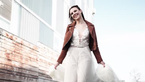 Very-lucky-and-happy-smiling-bride-in-brown-leather-jacket-walking-a-long-an-urban-environment-with-dress-in-her-hands