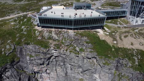 Spectacular-Loen-skylift-building-near-to-far-reverse-aerial---Roof-viewpoint-filled-with-tourists-at-sunny-summer-day---Dangerous-location-on-edce-of-cliff---Mountain-Hoven-in-Loen-Norway