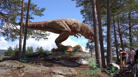 Tyrannosaurus-Rex-model-with-moving-body-parts---Massive-recreation-in-dinosauria-theme-park-Norway---Low-angle-looking-up-at-dinosaur-standing-on-hill-with-blue-sky-background-and-families-around