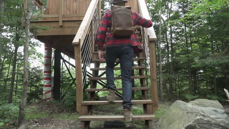 A-young-man-climbs-up-the-stairs-to-an-elevated-small-cabin-while-wearing-flannel-and-jeans-with-a-leather-back-in-slow-motion,-120-fps