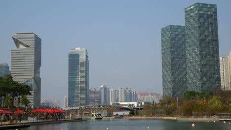 Moden-Urban-Skyline-of-Songdo-Central-Park,-G-Tower---People-Travel-by-Water-Taxi-Boat-on-a-Lake,-Travelers-Crossing-a-Footbridge