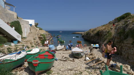 Barcos-Y-Gente-En-La-Orilla-Pedregosa-En-La-Playa-De-Port-Alga,-Polignano-A-Mare,-Apulia,-Italia
