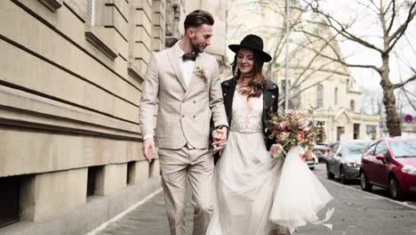 Retro-styled-wedding-couple-in-black-leather-jacket-and-hat-running-trough-the-city-streets-while-holding-hands