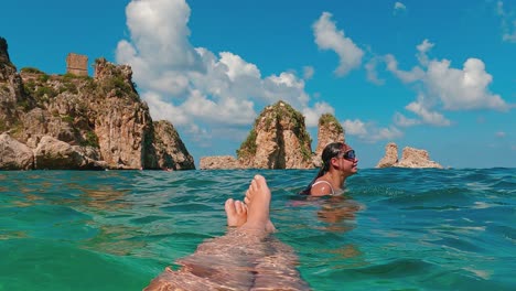 Fpv-of-male-legs-and-feet-relaxing-while-floating-on-sea-water-with-Scopello-Stacks-or-Faraglioni-watchtower-and-young-girl-with-diving-mask-in-background
