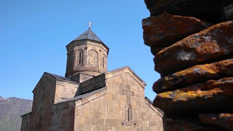 Dolly-Left-Reveal-Of-Gergeti-Church-Against-Clear-Blue-Skies-From-Pile-Of-Rocks