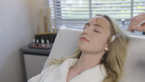 Young-blonde-caucasian-woman-receive-a-health-skin-care-facial-treatment-while-lying-on-a-massage-bed-in-luxury-resort-spa-saloon