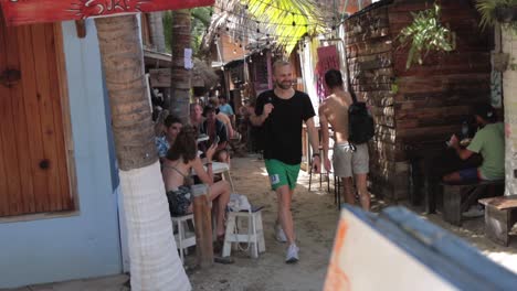 Male-shirtless-backpacker-almost-bumps-into-a-tourist-walking-in-same-direction-near-an-outdoor-hippy-food-court-with-muddy-dirt-roads-and-cozy-shacks,-restaurants-and-bars