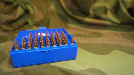 Ammunition-box-full-of-powerful-military-grade-223-caliber-varmint-bullets-with-blue-plastic-tip-laying-on-camouflage-jacket---Slider-from-left-to-right