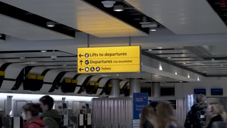 Yellow-directional-signage-at-Heathrow-International-Airport-Terminal-3,-passengers-passing-by-going-about-their-business-making-their-way-to-the-departure’s-terminal,-London,-England