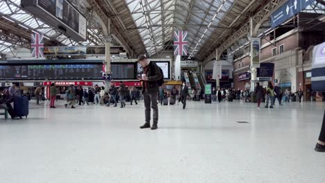 Inside-View-Of-London-Victoria-Station-With-Adult-Male-Checking-His-Phone-And-Commuters-Walking-In-Background