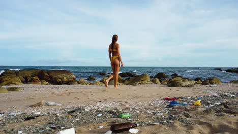 Tan-blonde-model-in-bikini-walking-alone-on-dirty-polluted-beach-with-trash