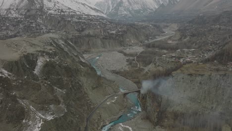 Luftaufnahme-über-Dem-Türkisfarbenen-Fluss-Im-Hunza-Tal-Mit-Rauch,-Der-In-Der-Nähe-Der-Brücke-Aufsteigt