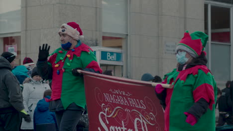 Two-people-in-elf-costumes-holding-Santa-Claus-parade-banner-in-Christmas-Parade,-Niagara-Falls,-Ontario