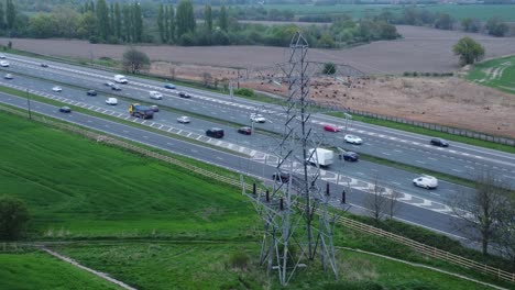 Vehicles-on-M62-motorway-passing-pylon-tower-on-countryside-farmland-fields-aerial-descending-view