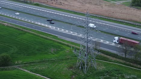 Vehicles-on-M62-motorway-passing-pylon-tower-on-countryside-farmland-fields-aerial-view-orbit-right