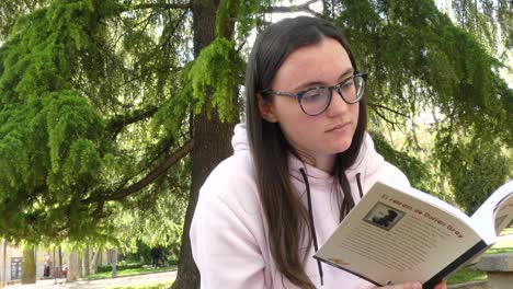 Student-girl-reading-a-novel-next-to-a-tree-on-a-park,-outdoors-video