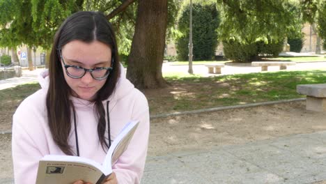 Cute-girl-reading-a-novel-on-the-street,-next-to-a-park