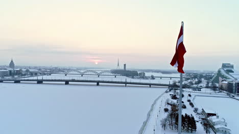 Luftaufnahme-Der-Hoch-Fliegenden-Lettischen-Flagge-Mit-Blick-Auf-Den-Zugefrorenen-Fluss-Und-Die-Schneebedeckten-Ufer-Eines-Eisbedeckten-Flusses-Im-Hintergrund