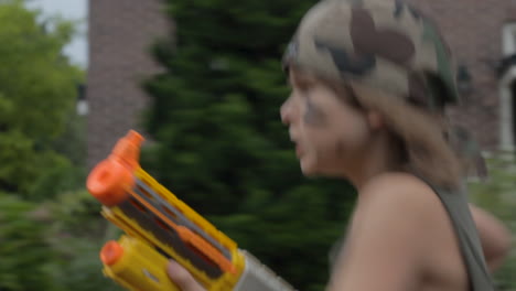 Young-boy-playing-soldier-with-toy-gun-running-through-beautiful-suburban-neighborhood