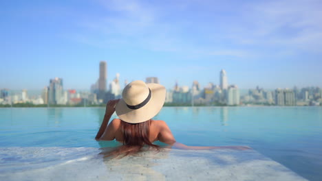 Lonesome-Woman-With-Floppy-Hat-in-Rooftop-Infinity-Pool-With-View-of-Urban-Sunny-Cityscape,-Back-View