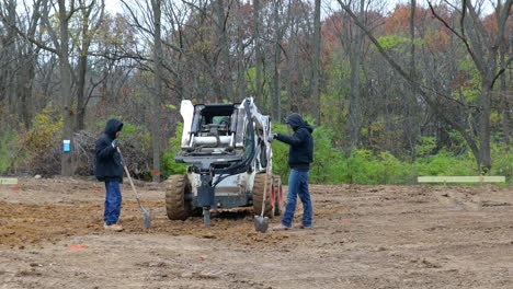 Three-construction-workers-using-shovels-and-hydraulic-auger-mounted-on-skid-steer-loader-to-dig-a-post-hole-for-the-frame-of-new-barn-construction