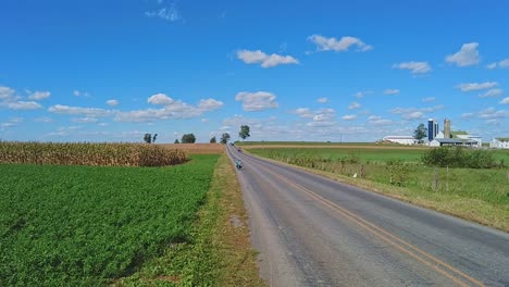 Un-Niño-Amish-Montando-Una-Moto,-En-Una-Posición-Agachada,-Bajando-Por-Una-Colina-De-Carretera-Rural,-En-Un-Día-Soleado