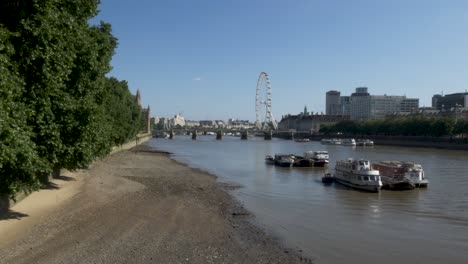 Aufnahme-Mehrerer-Festgemachter-Boote-Entlang-Der-Themse-Bei-Ebbe,-In-Der-Ferne-Das-Ikonische-Riesenrad-London-Eye-Und-Die-Westminster-Bridge-An-Einem-Schönen-Sonnigen-Tag,-London,-England