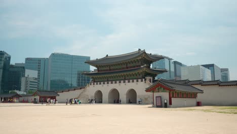 Turistas-Caminando-Por-La-Puerta-Gwanghwamun-Hasta-La-Plaza-Del-Palacio-Gyeongbokgung-Con-Edificios-De-Oficinas-De-Gran-Altura-De-Negocios-De-Seúl-Contra-El-Cielo-Azul---Vista-Lejana