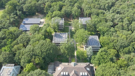 Drone-flying-away-from-several-small-office-buildings-with-solar-panels-on-rooftop-in-a-beautiful-green-forest