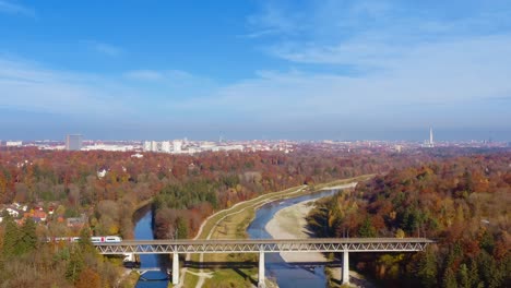 Charming-travel-clip---flying-by-drone-at-the-autumn-season-over-the-Isar-river,-a-train-is-passing-by-a-bridge-with-the-look-at-the-popular-tourism-city-of-Munich-in-southern-germany-under-blue-sky