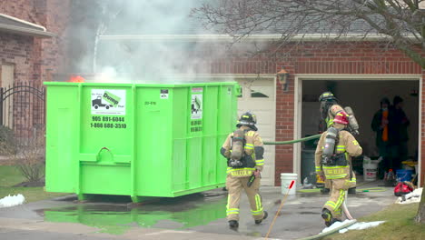 Ein-Feuerwehrauto-Fährt-Vorbei,-Während-Feuerwehrleute-Am-Brandort-Eines-Brennenden-Müllcontainers-Eingreifen