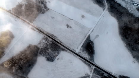 Aerial-drone-flying-above-a-road-in-the-middle-of-a-vast-snow-covered-field-on-a-cold-winter-morning-with-cars-passing-by-on-the-road-on-a-cloudy-day