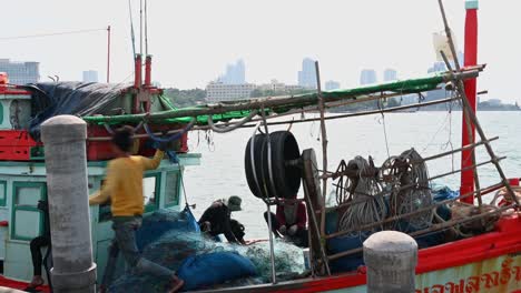 A-fisherman-stands-up-to-go-to-the-side-of-the-boat-while-others-are-mending-nets,-Pattaya-Fishing-Dock,-Chonburi,-Thailand